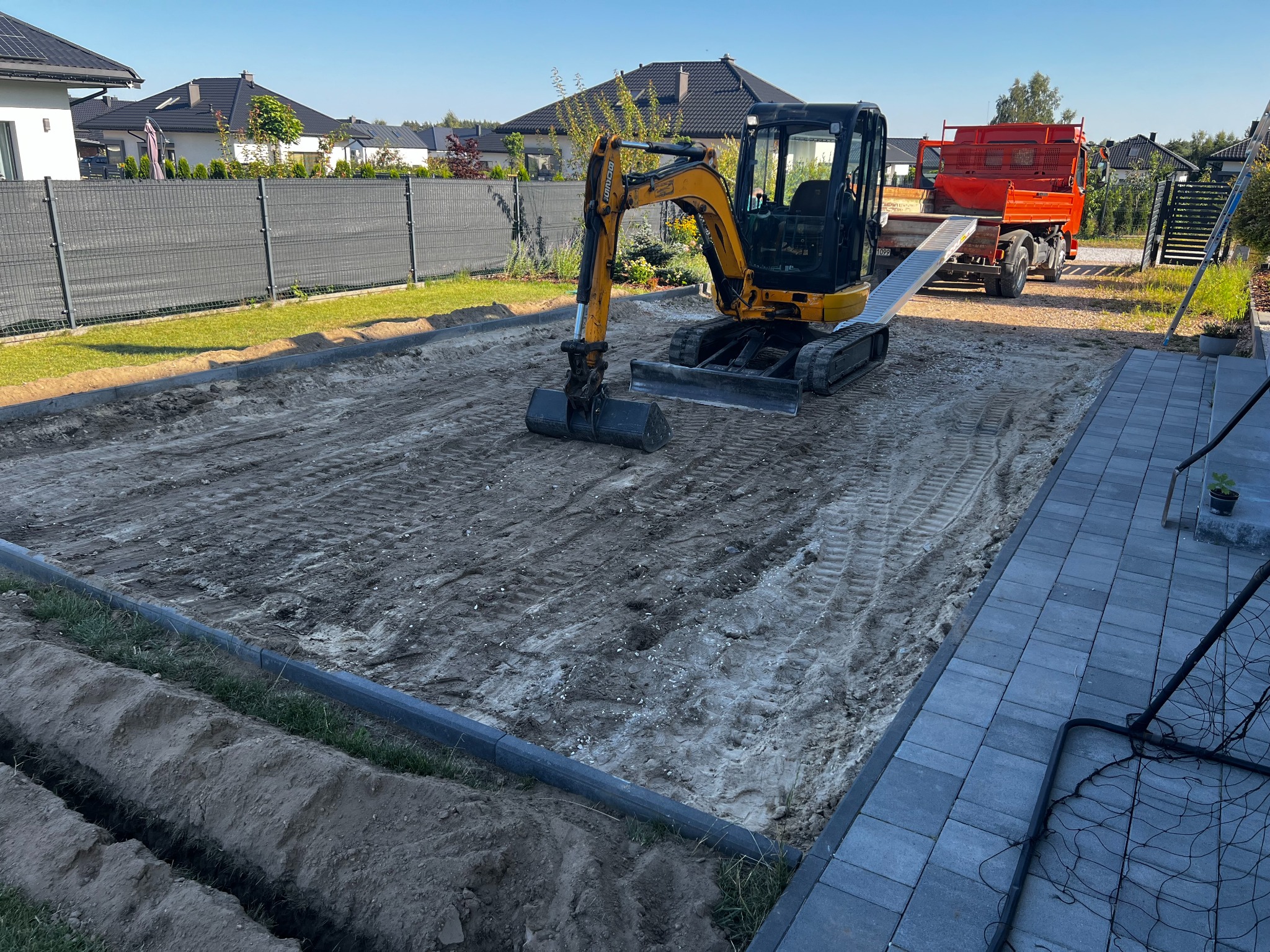 Żółta mini koparka na terenie przygotowanym pod kostkę brukową, z widocznymi śladami opon i wykopem pod obrzeża. W tle domy jednorodzinne i transportowa ciężarówka.