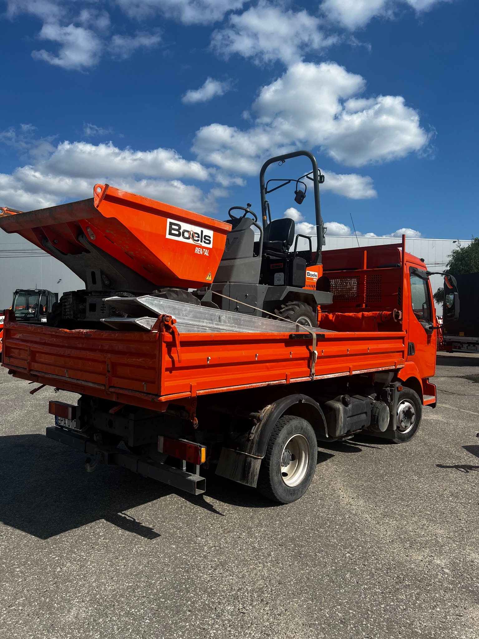 Pomarańczowa ciężarówka z małą koparką Boels na pace, zaparkowana na placu, błękitne niebo w tle. Widok z tyłu, skupienie na maszynach i tablicy rejestracyjnej.