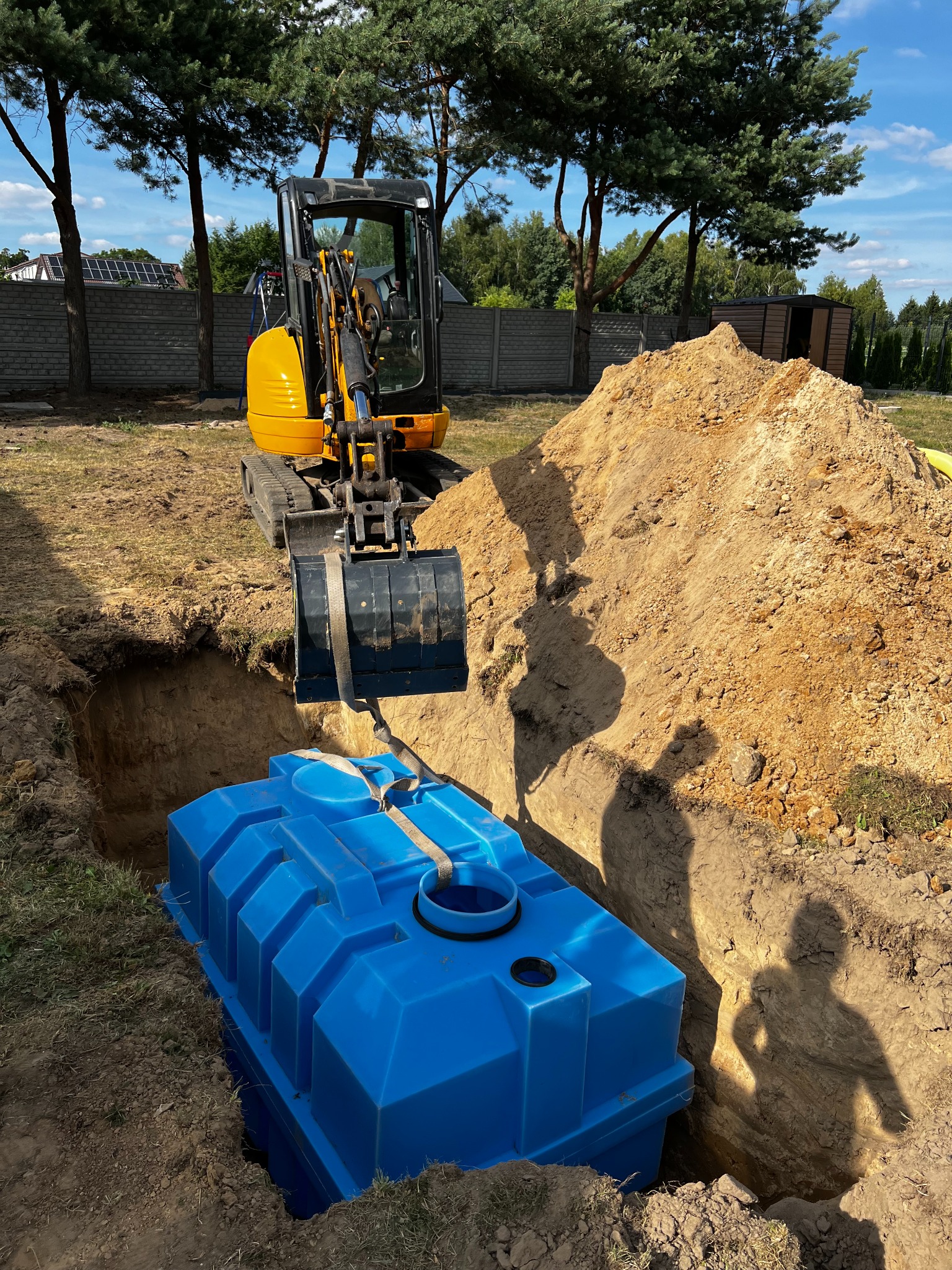 Żółta mini koparka umieszcza niebieski, plastikowy zbiornik w wykopie. W tle drzewa, ogrodzenie i dom z panelami słonecznymi. Wykopane ziemia tworzy kopiec.