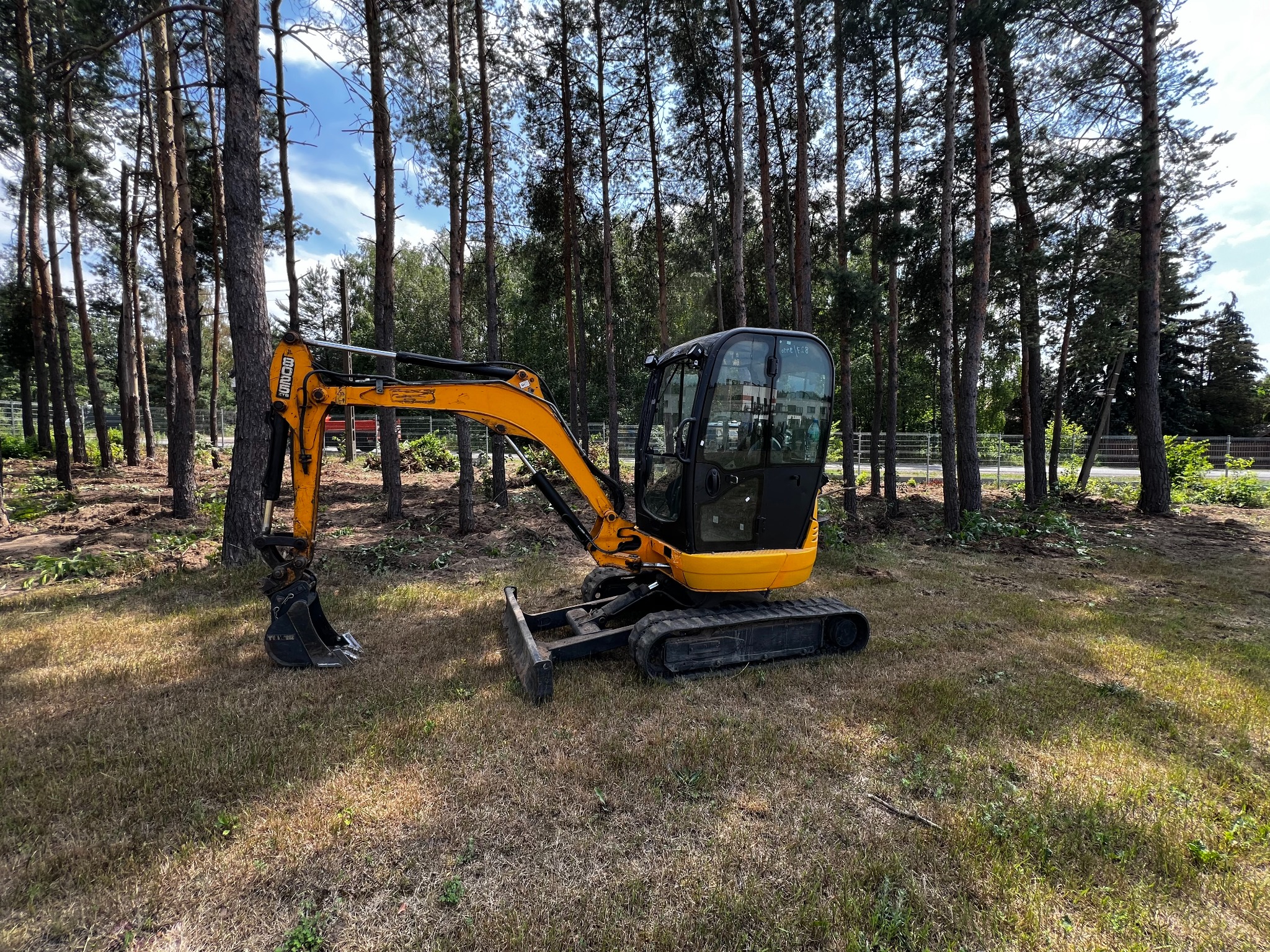Żółta mini koparka JCB na tle lasu sosnowego, gotowa do pracy przy wykopie pod instalacje. Widoczne ślady po wycince krzewów i drzew.