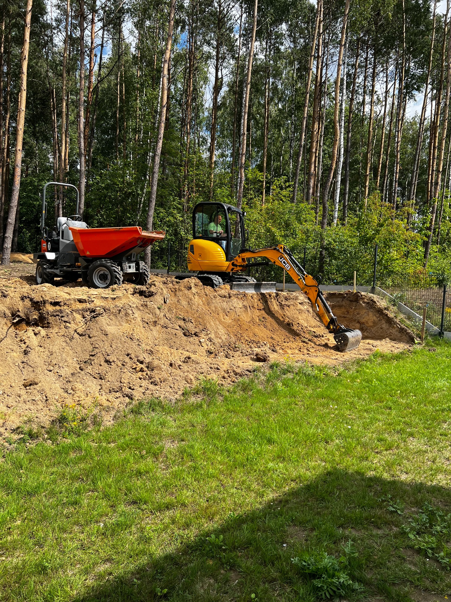 Żółta mini koparka JCB i wywrotka na wykopanym terenie. W tle wysokie drzewa, częściowo ogrodzenie. Prace ziemne w toku, widoczny wykop.