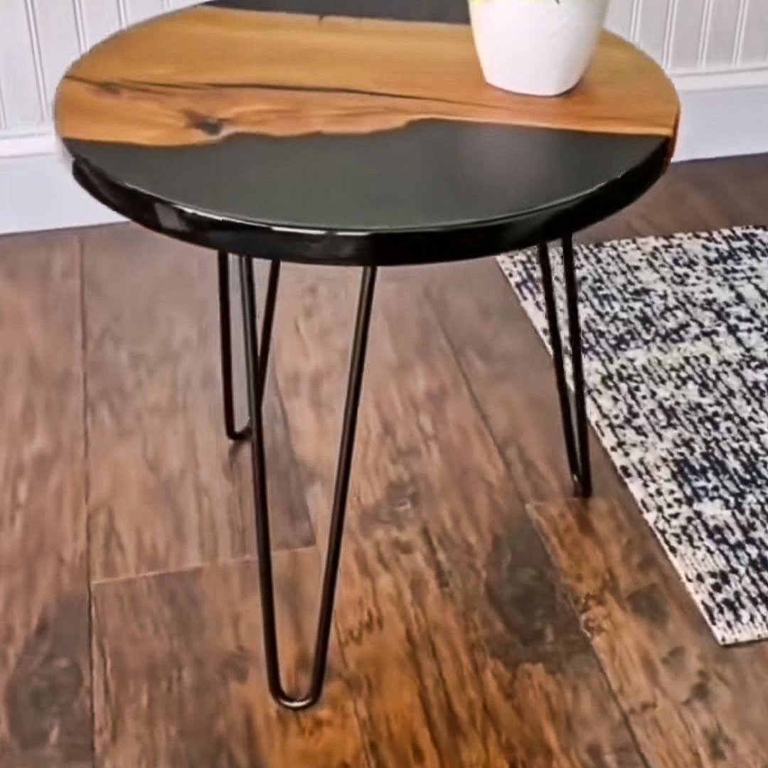 Okrągły stolik kawowy z blatem z drewna i czarnej żywicy epoksydowej, osadzony na trzech metalowych nogach w stylu hairpin, na tle drewnianej podłogi i fragmentu dywanu.