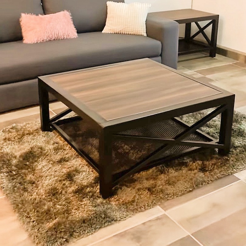 Kwadratowy stolik kawowy z drewnianym blatem i metalową ramą z krzyżowymi wzmocnieniami, ustawiony na puszystym dywanie w salonie z szarą sofą i dekoracyjnymi poduszkami.