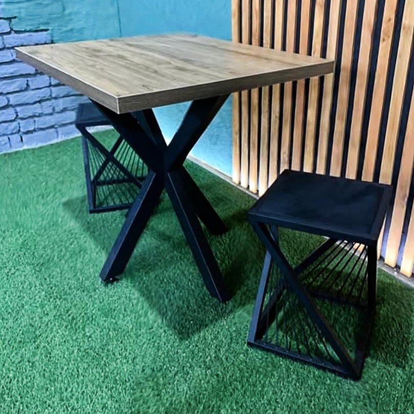 Kwadratowy stół z blatem imitującym drewno i czarną, metalową podstawą w kształcie litery X, w komplecie z dwoma metalowymi stołkami o geometrycznej konstrukcji, ustawiony na sztucznej trawie...