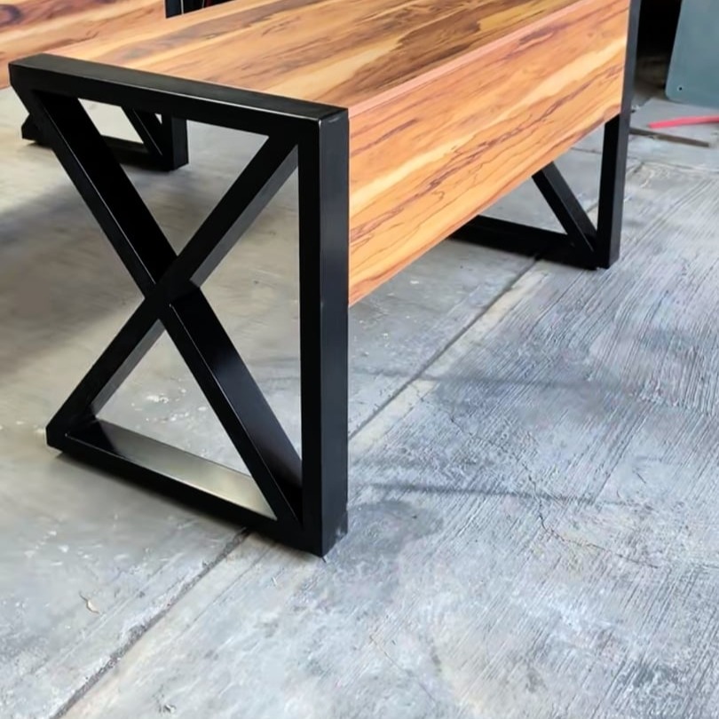 Stolik kawowy z blatem z egzotycznego drewna i czarną, metalową podstawą w kształcie litery X, widoczny na tle betonowej posadzki.