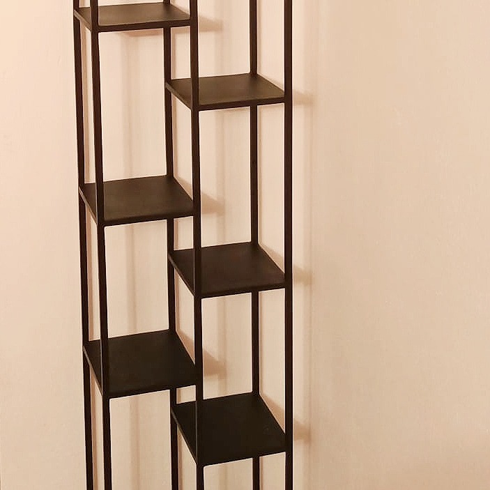 Czarny, metalowy regał o asymetrycznej konstrukcji, z kwadratowymi półkami, ustawiony na tle jasnej ściany.