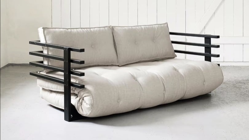Beżowa sofa futon z czarnymi metalowymi podłokietnikami w minimalistycznym wnętrzu.