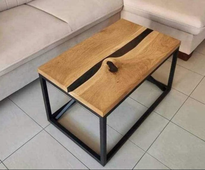 Stolik kawowy z blatem z jasnego drewna z żywicą epoksydową w kolorze czarnym, osadzony na czarnej, metalowej ramie, na tle jasnej sofy i płytek podłogowych.