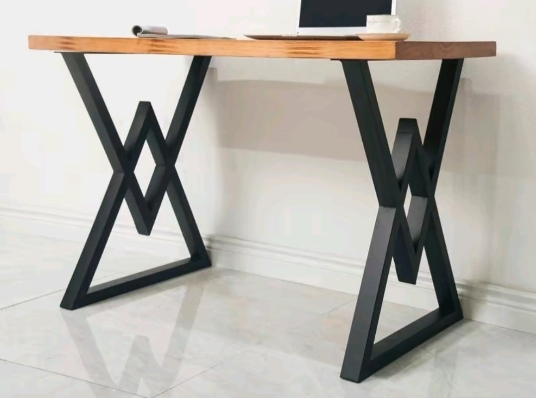 Stół z blatem z jasnego drewna i czarnymi, geometrycznymi nogami w kształcie litery X z dodatkowymi elementami, na blacie laptop i filiżanka.