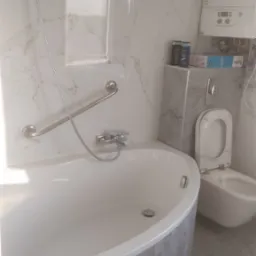 Nowoczesna łazienka z wanną obudowaną szarymi płytkami imitującymi marmur, wiszącą toaletą z bidetem, chromowaną armaturą i grzejnikiem Junkers na ścianie wyłożonej białym marmurem.