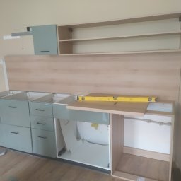 Ryszard Kowalewski - Montaż dolnych i górnych szafek kuchennych w kolorze pastelowej zieleni, z poziomnicą na blacie, widoczne elementy konstrukcyjne i mocowania, w trakcie instalacji.