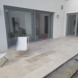 Jasne płytki tarasowe ułożone na zewnątrz budynku, z dwoma białymi leżakami i widocznym obszarem przygotowanym pod kolejne płytki, ukazującym proces układania.