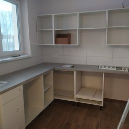 Ryszard Kowalewski - Montaż białych szafek kuchennych z szarym blatem, widoczna poziomica, drabina i karton w szafce, okno z widokiem na drzewa.