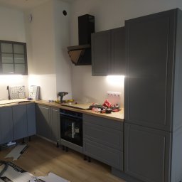 Ryszard Kowalewski - Montaż szarej kuchni z drewnianym blatem: widoczne narzędzia, płyty podłogowe i niedokończone prace wykończeniowe.