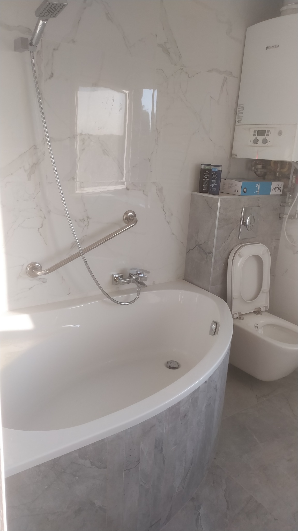 Nowoczesna łazienka z wanną obudowaną szarymi płytkami imitującymi marmur, wiszącą toaletą z bidetem, chromowaną armaturą i grzejnikiem Junkers na ścianie wyłożonej białym marmurem.