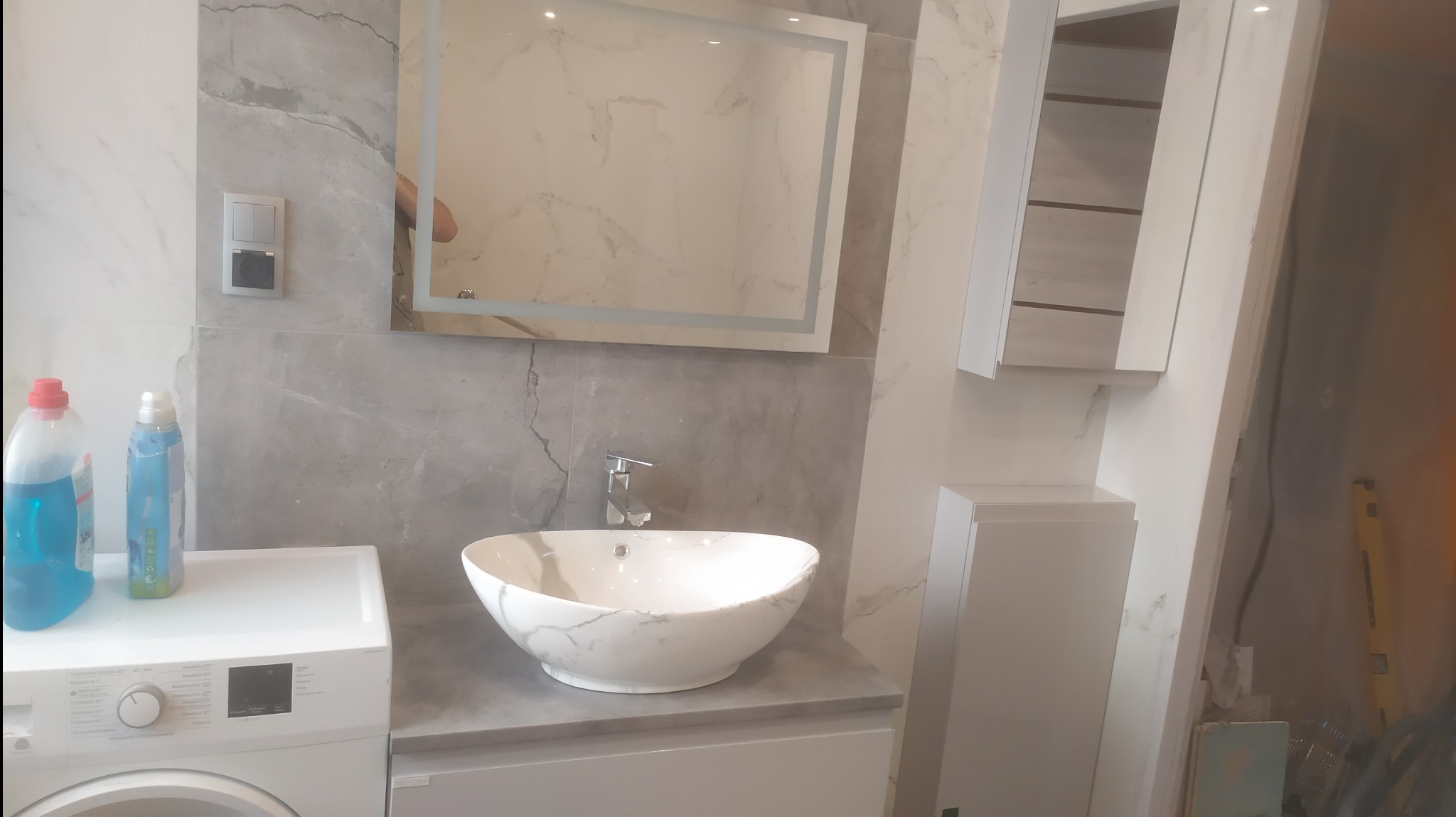 Nowoczesna łazienka z umywalką nablatową w marmurowym wzorze, podświetlanym lustrem i pralką z płynami do prania po lewej stronie, w trakcie wykańczania.