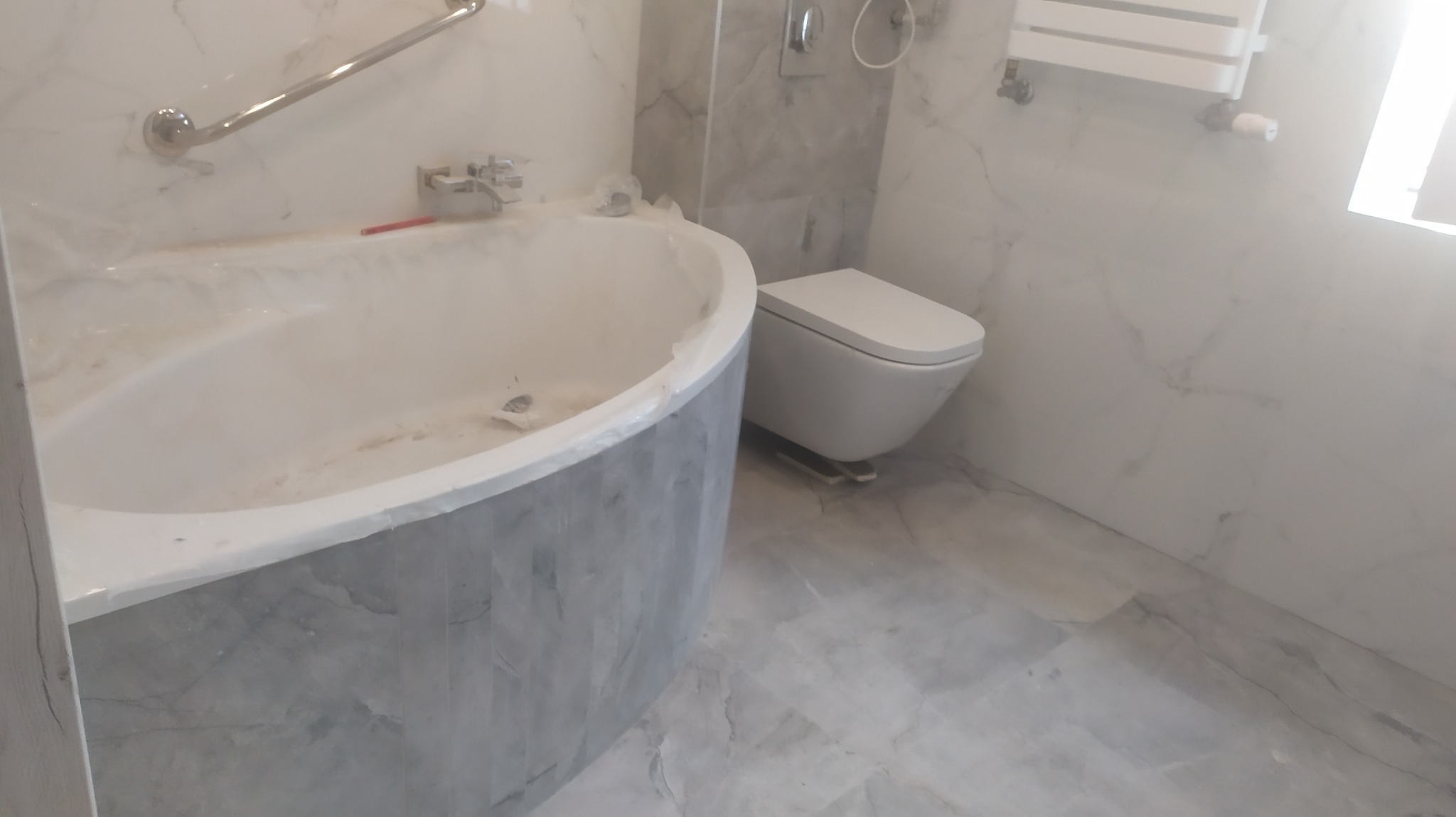 Nowoczesna łazienka z owalną wanną z szarą obudową imitującą marmur, wiszącą toaletą i grzejnikiem drabinkowym na ścianie wyłożonej płytkami imitującymi biały marmur z szarymi żyłami.