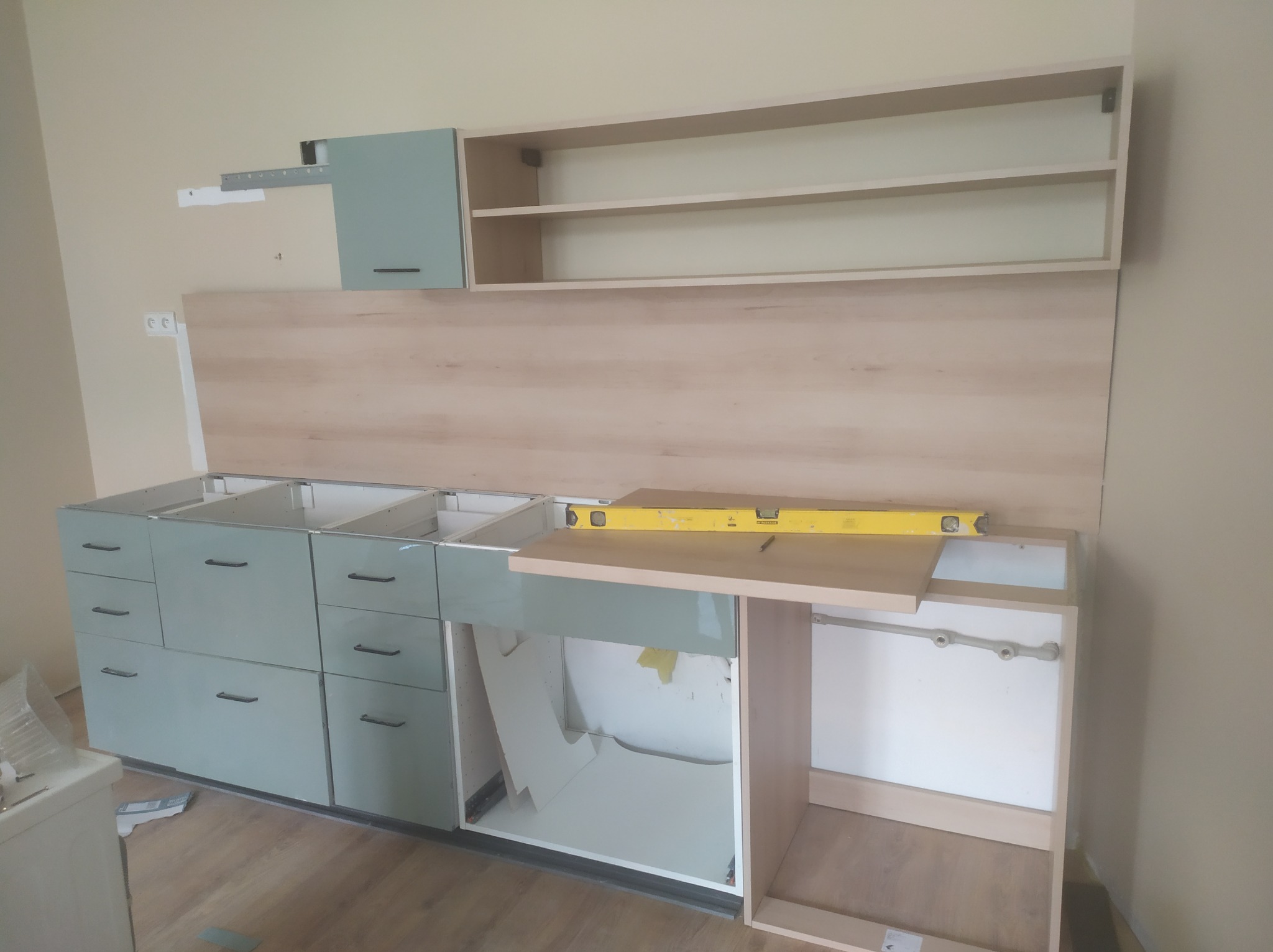 Montaż dolnych i górnych szafek kuchennych w kolorze pastelowej zieleni, z poziomnicą na blacie, widoczne elementy konstrukcyjne i mocowania, w trakcie instalacji.