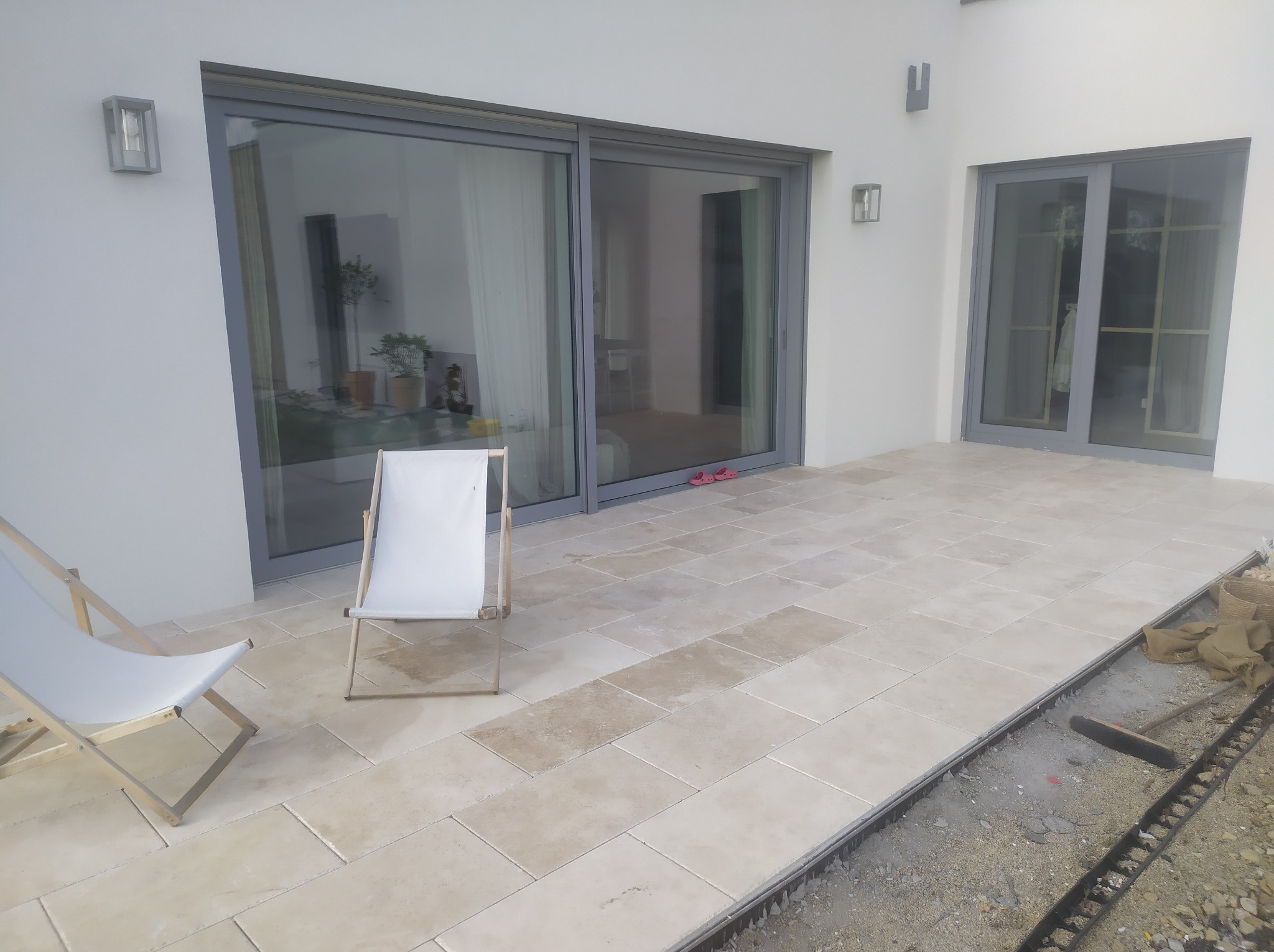 Jasne płytki tarasowe ułożone na zewnątrz budynku, z dwoma białymi leżakami i widocznym obszarem przygotowanym pod kolejne płytki, ukazującym proces układania.