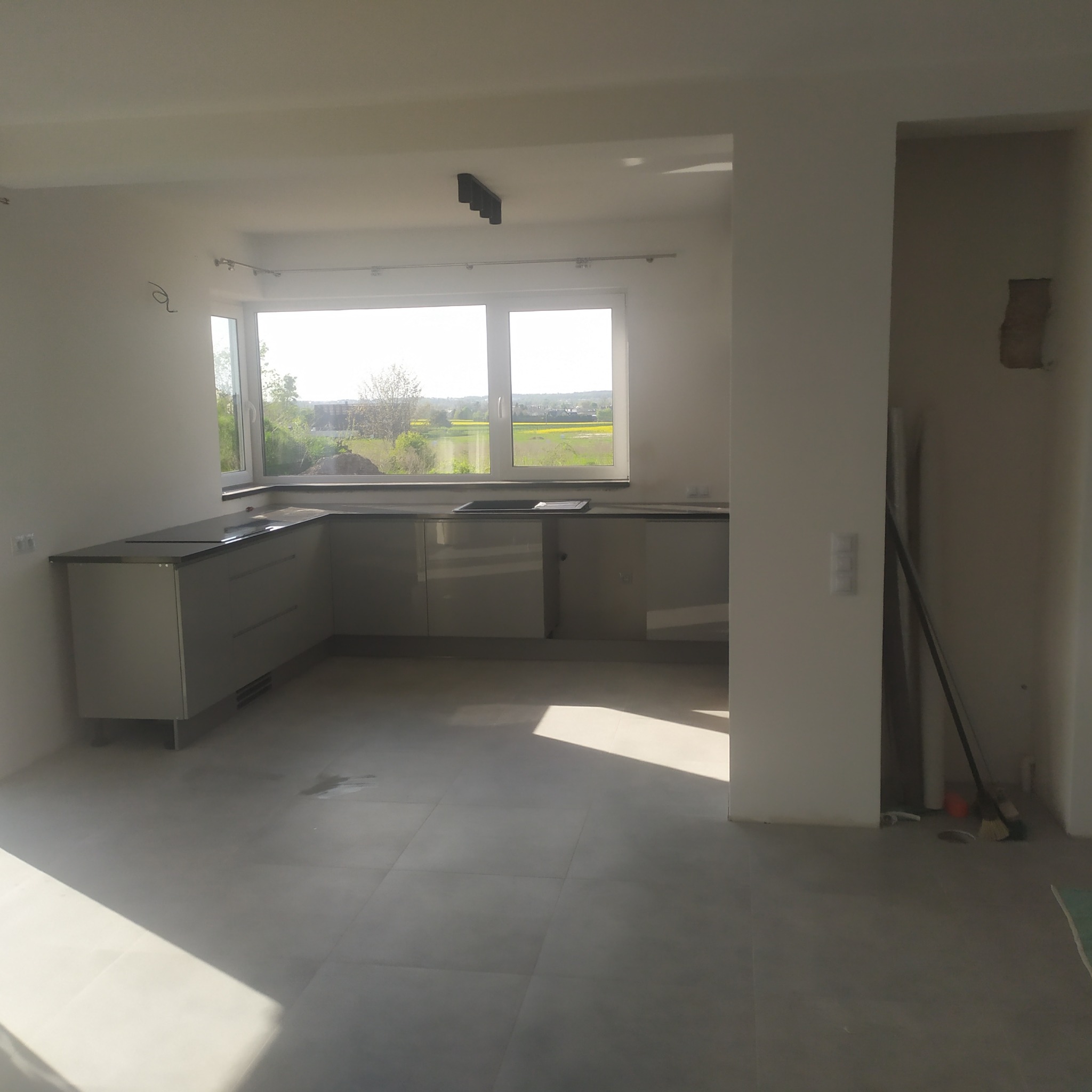 Jasna, minimalistyczna kuchnia z szarymi szafkami i blatem, widok na zielone pola za oknem; widoczne niedociągnięcia w wykończeniu ścian i podłogi.