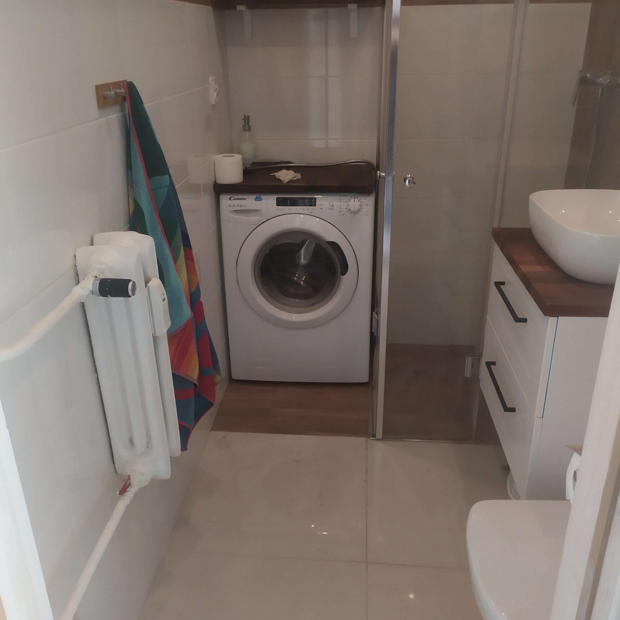Wnętrze łazienki z pralką Candy umieszczoną pod blatem, białą umywalką nablatową na szafce z ciemnym blatem i białym grzejnikiem żeberkowym z kolorowym ręcznikiem.