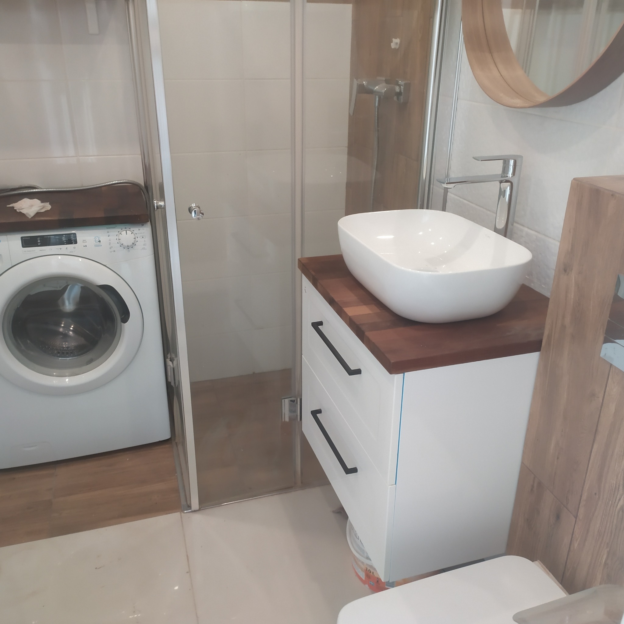 Biała umywalka nablatowa o zaokrąglonym kształcie na szafce z dwoma szufladami z ciemnym blatem w łazience z kabiną prysznicową i pralką.