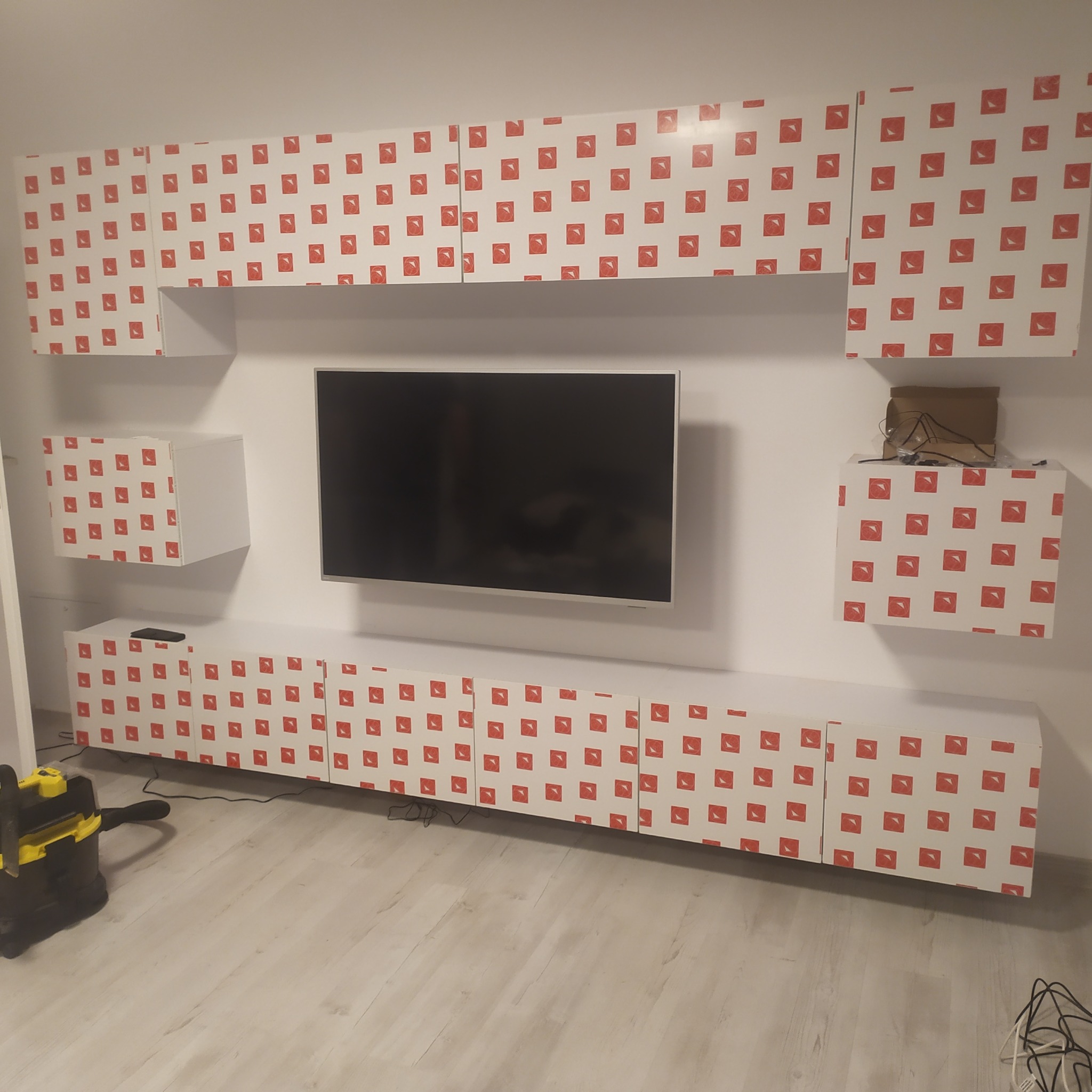 Biała meblościanka z czerwonym wzorem kwadratów, zawieszona na ścianie nad telewizorem; odkurzacz przemysłowy na podłodze sugeruje prace wykończeniowe w toku.