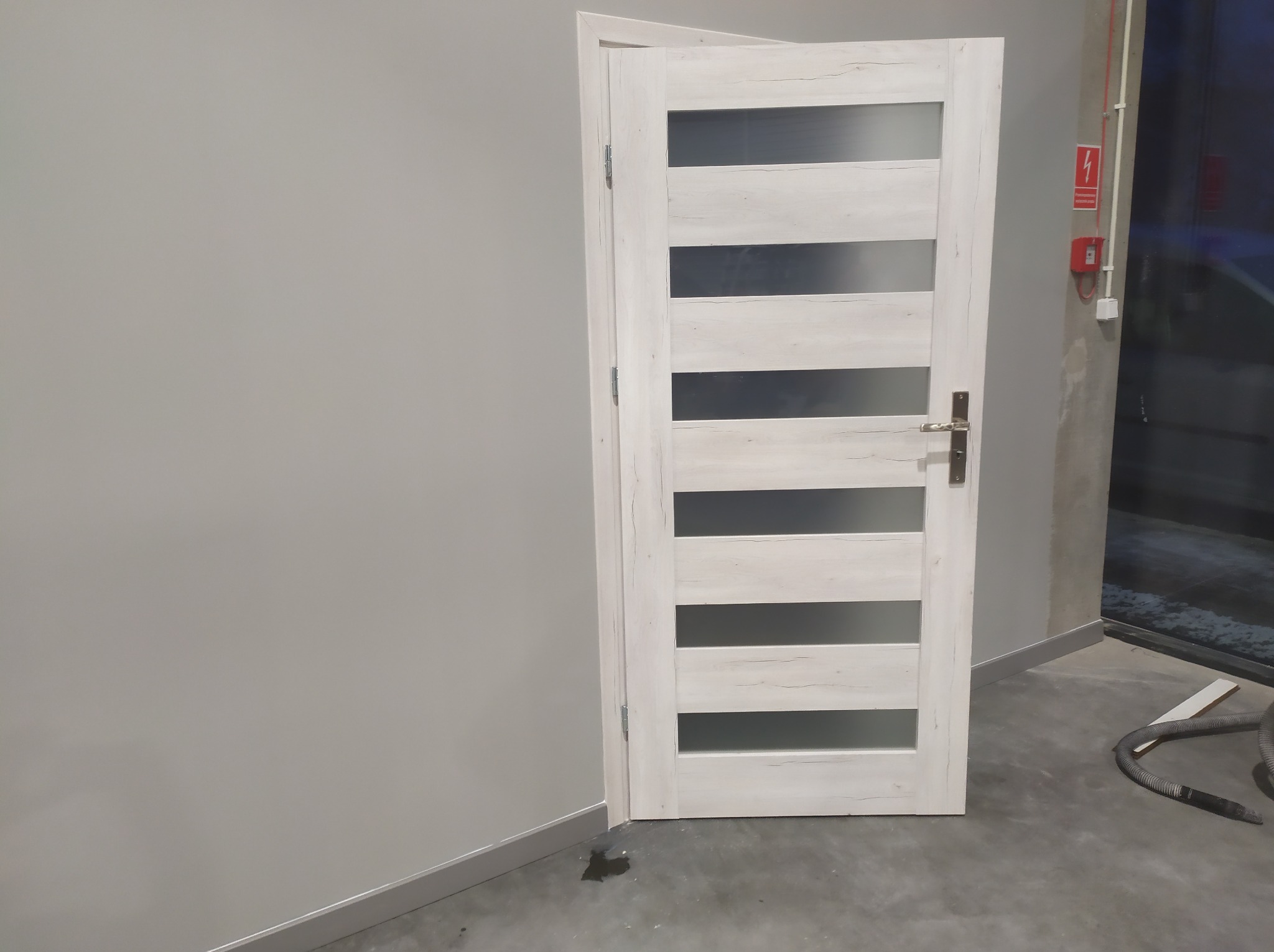 Zamknięte, białe drzwi wewnętrzne z przeszkleniami w nowym, szarym pomieszczeniu, z widocznym alarmem przeciwpożarowym i wężem odkurzacza na podłodze.