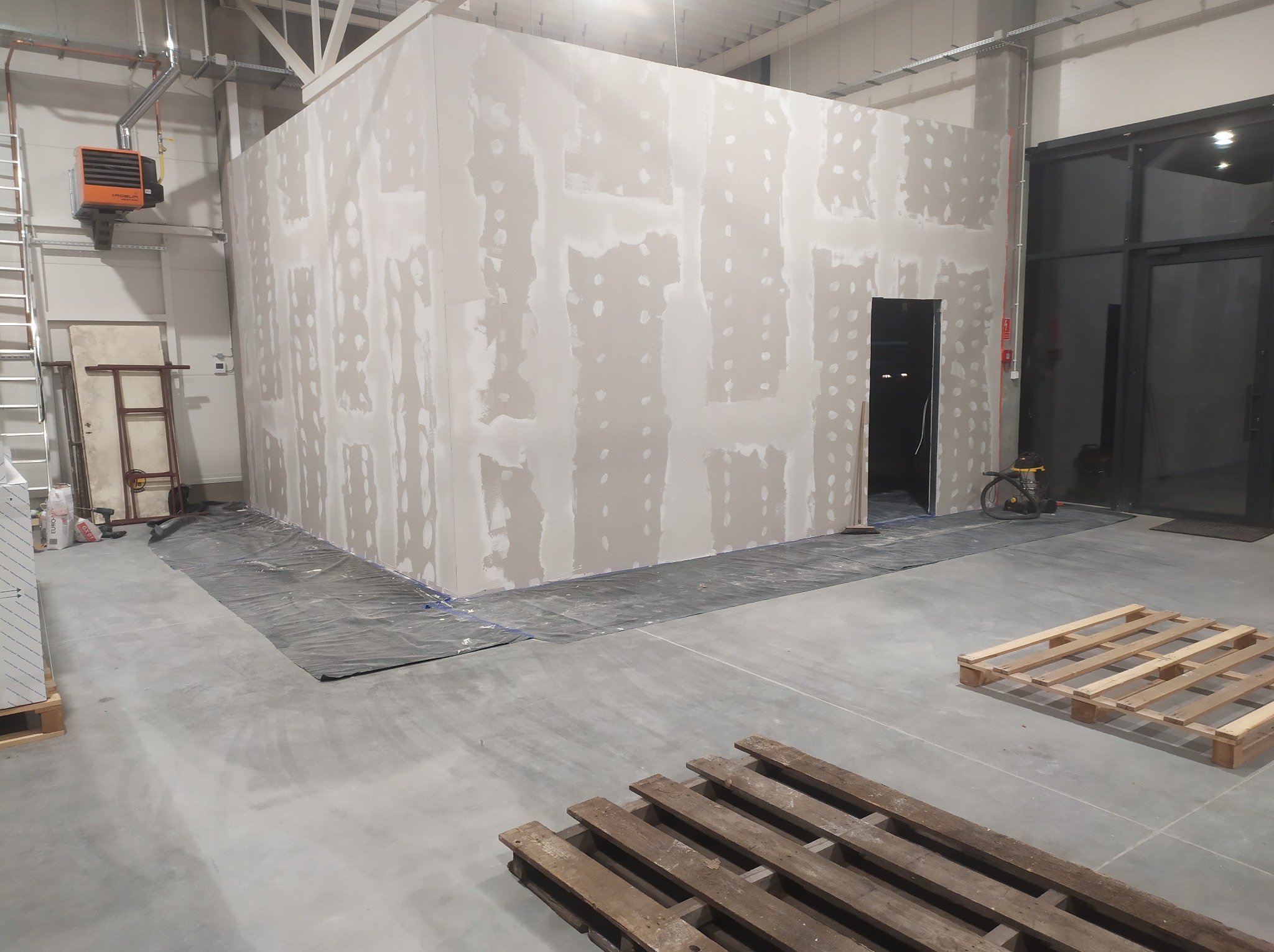 Wnętrze hali w trakcie budowy ścian działowych z płyt gipsowo-kartonowych, widoczne szpachlowanie łączeń płyt, podłoga zabezpieczona folią, palety na posadzce.