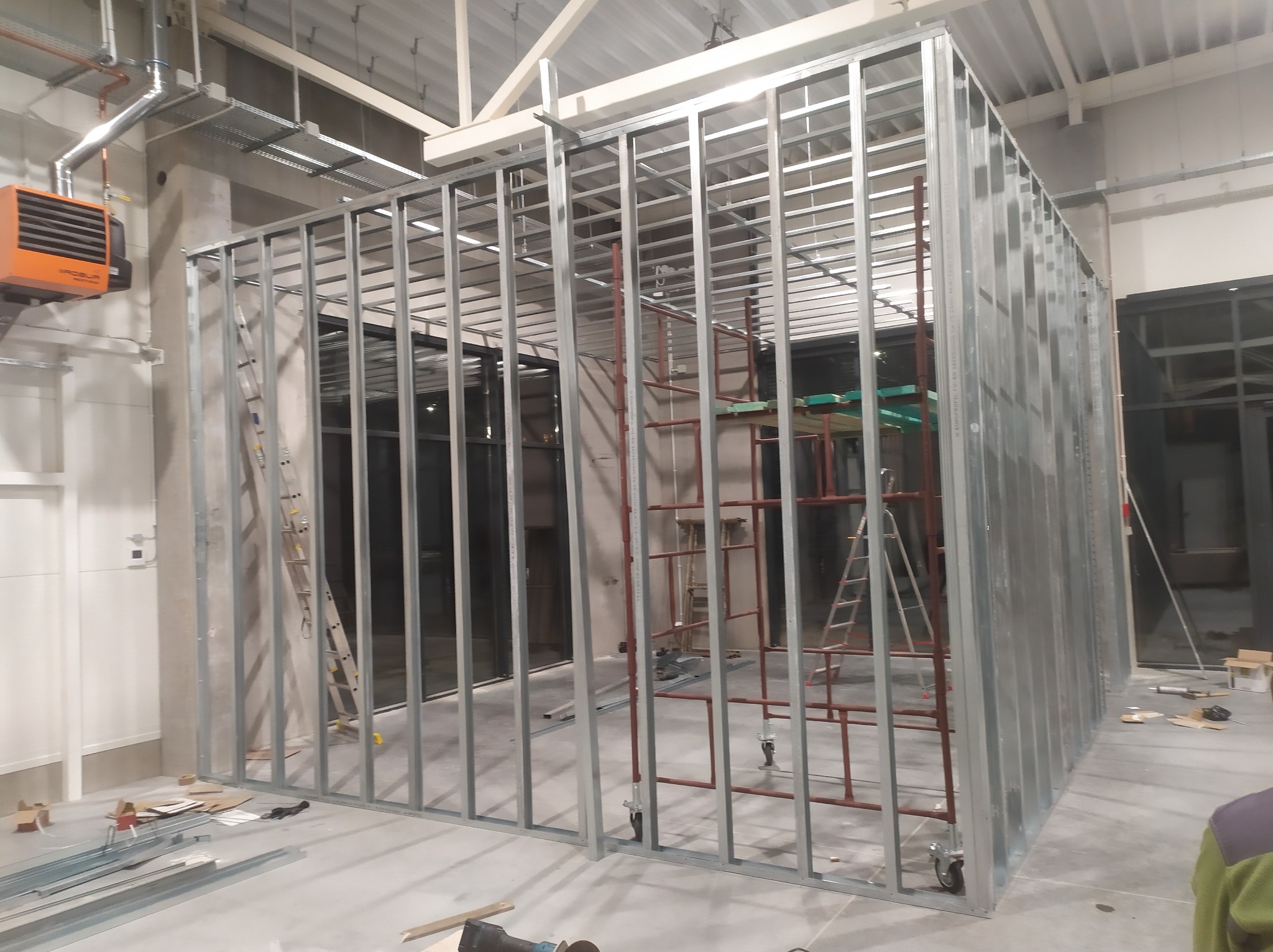 Metalowa konstrukcja ścian działowych w surowym stanie, widoczne drabiny i narzędzia na podłodze, pomarańczowy grzejnik na ścianie w tle.