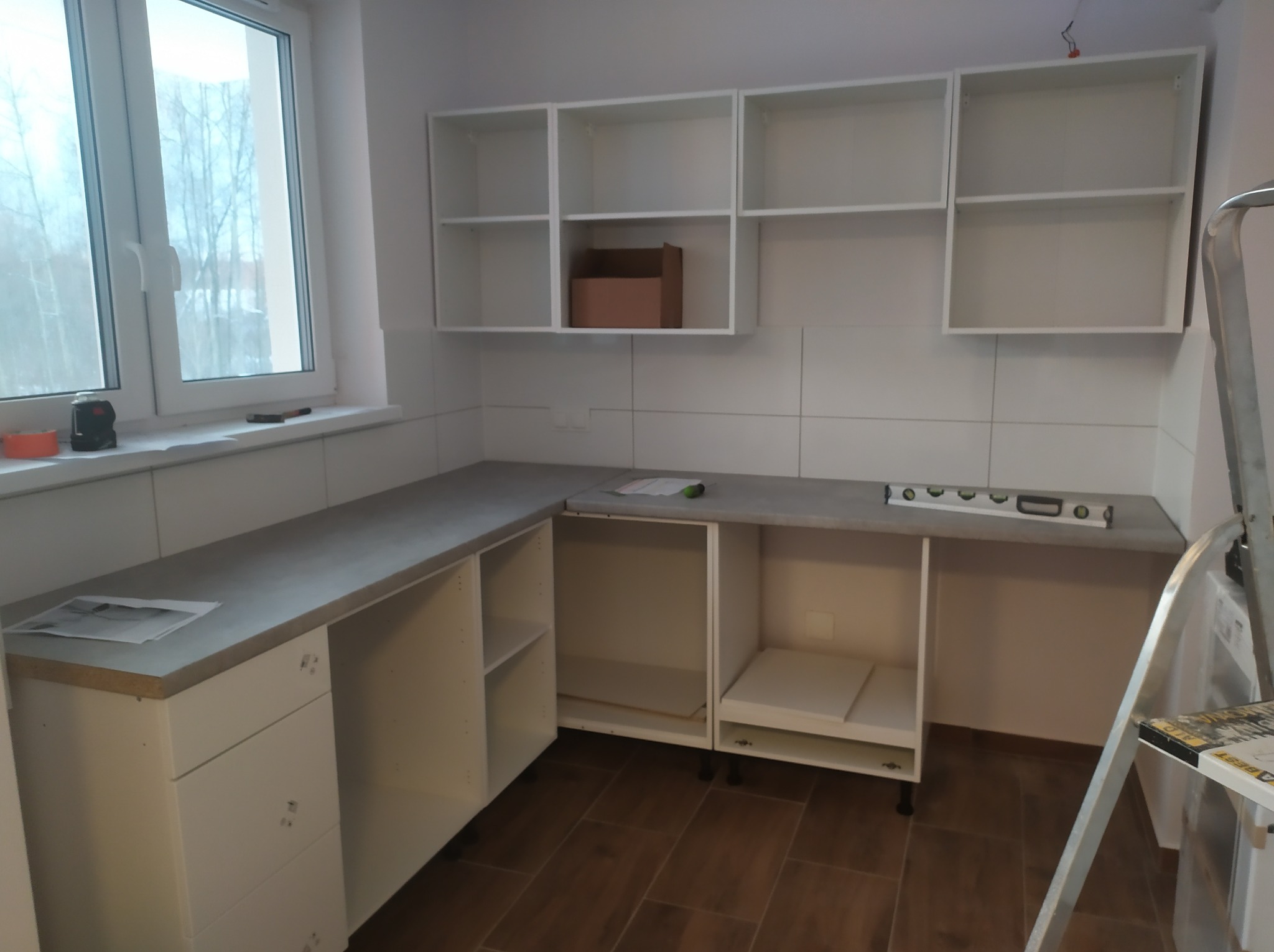 Montaż białych szafek kuchennych z szarym blatem, widoczna poziomica, drabina i karton w szafce, okno z widokiem na drzewa.
