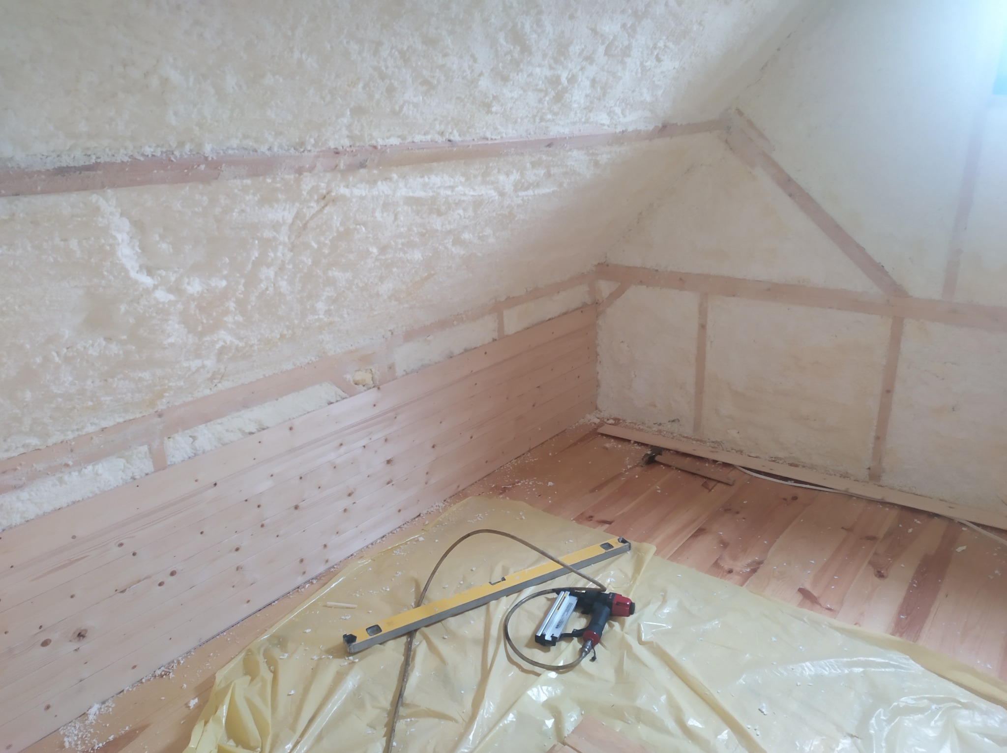 Poddasze w trakcie ocieplania pianką, widoczne drewniane panele boazeryjne i narzędzia pracy: poziomica i pistolet do gwoździ, zabezpieczone folią malarską.