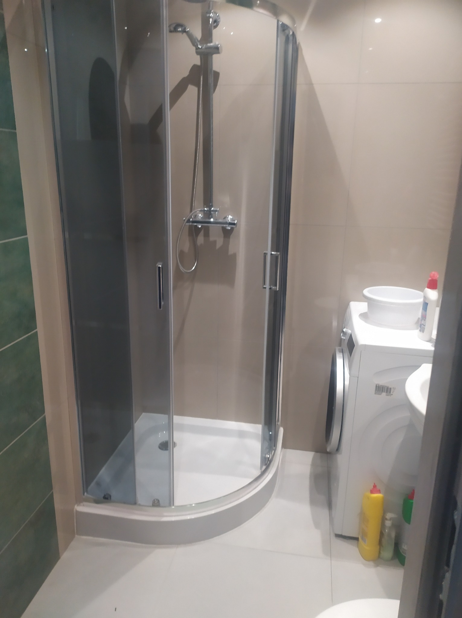 Narożna kabina prysznicowa z chromowaną armaturą, umieszczona w łazience z beżowymi płytkami, obok pralki z miską i środkami czystości.