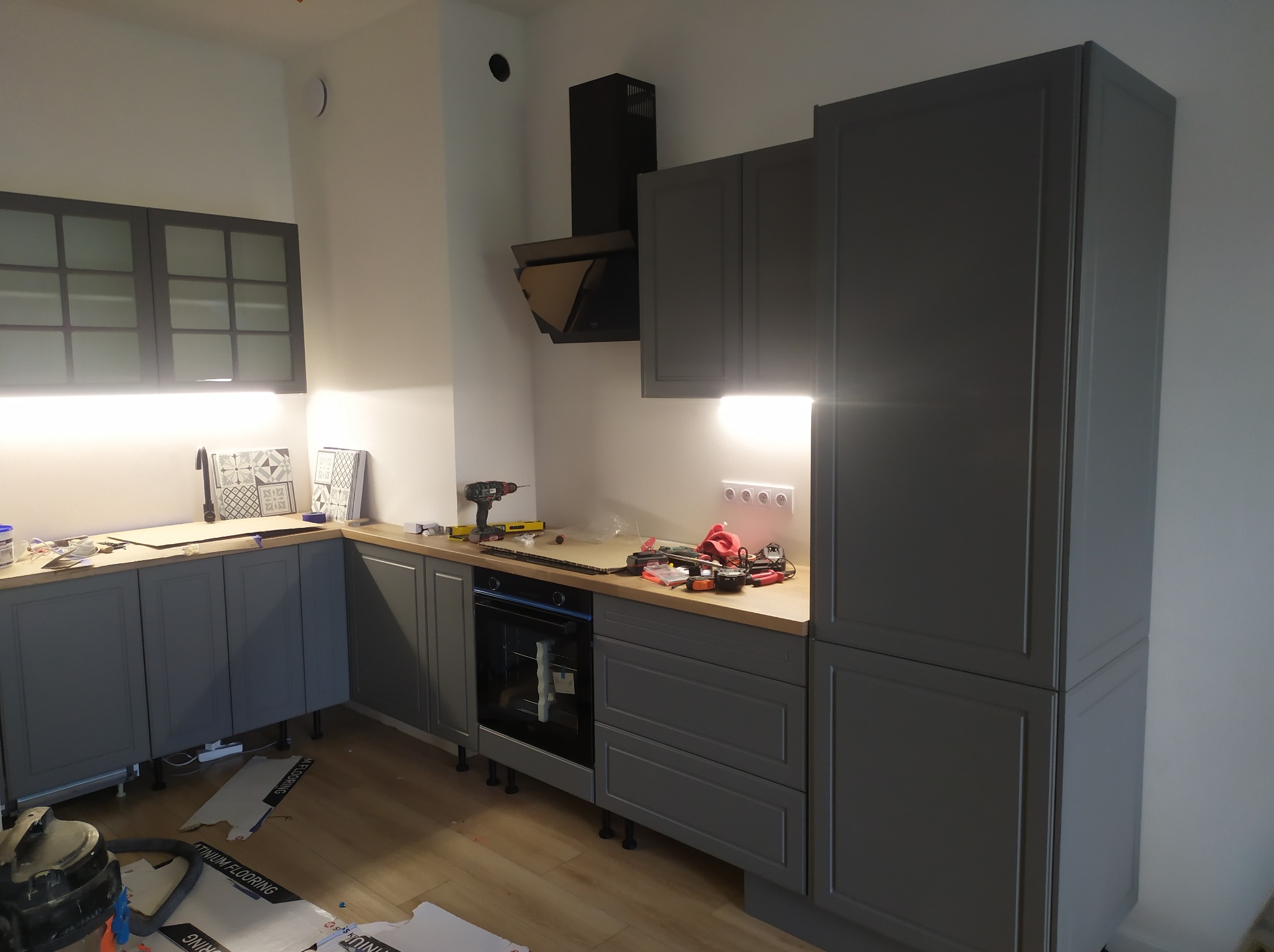 Montaż szarej kuchni z drewnianym blatem: widoczne narzędzia, płyty podłogowe i niedokończone prace wykończeniowe.