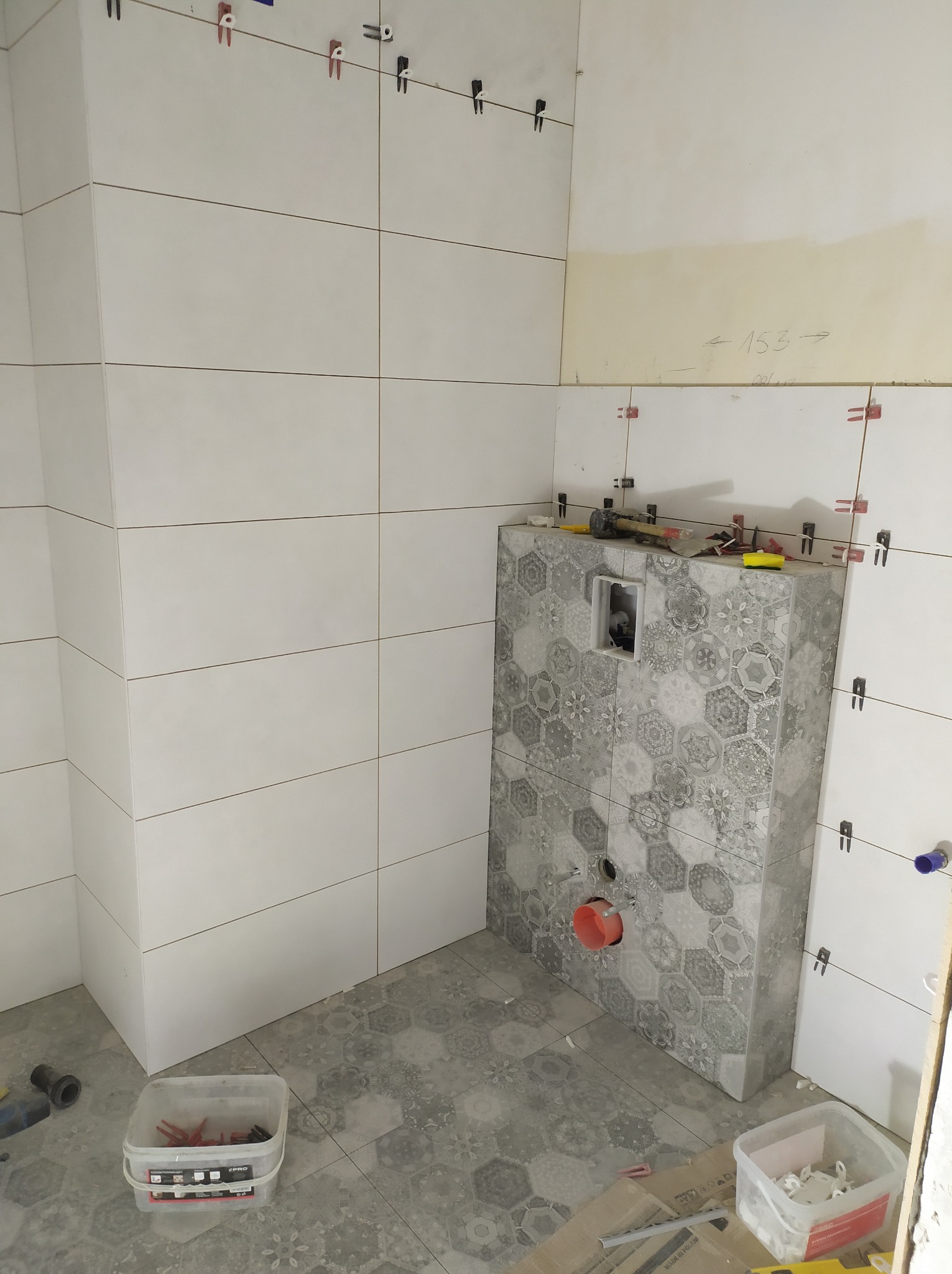 Montaż płytek w łazience w trakcie realizacji: białe, prostokątne płytki ścienne, heksagonalne płytki podłogowe i na zabudowie stelaża WC, widoczne narzędzia i akcesoria glazurnicze.