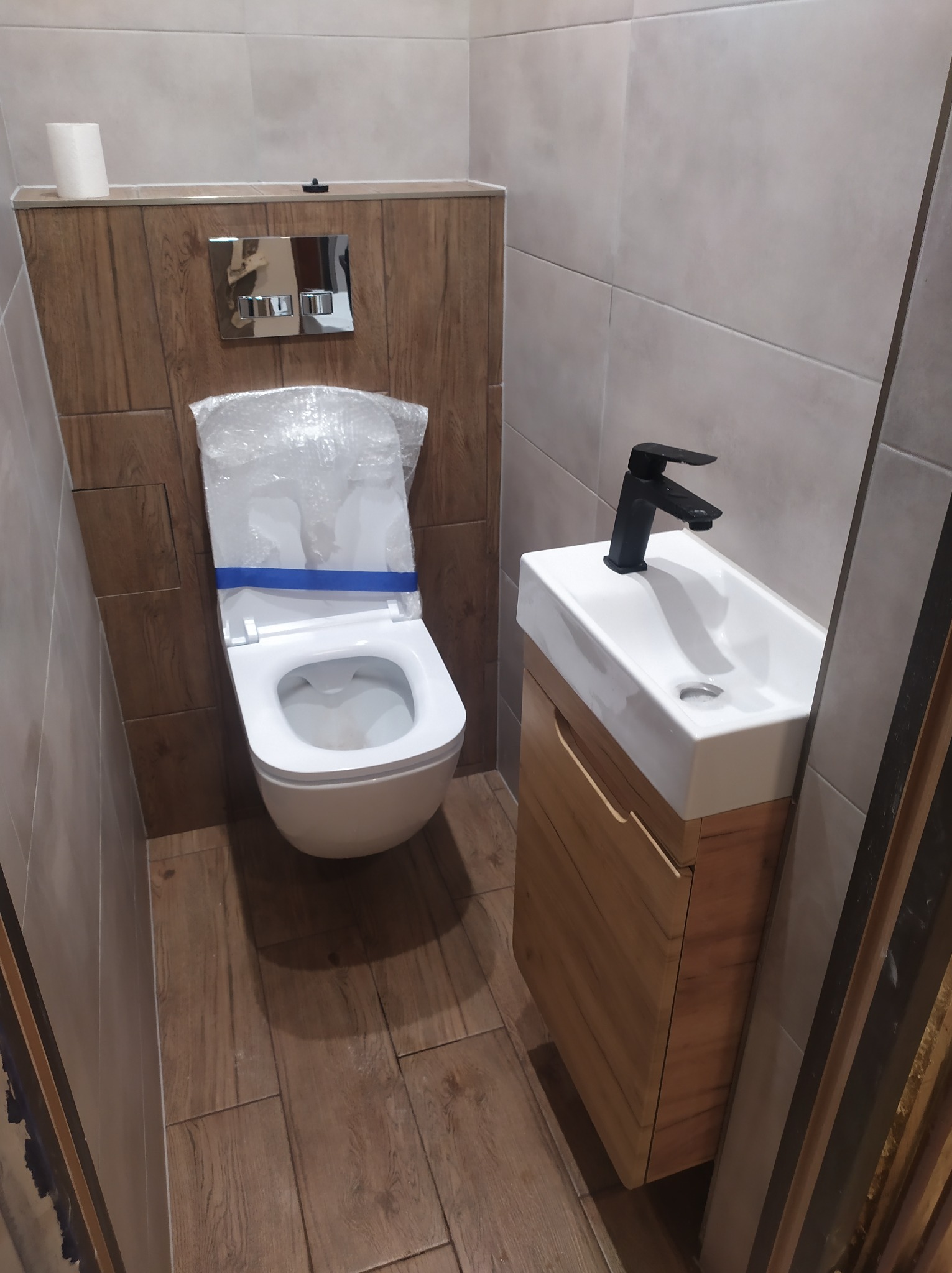 Kompaktowa łazienka z podwieszaną toaletą zabezpieczoną folią i małą umywalką z czarną baterią, wykończona płytkami imitującymi drewno.