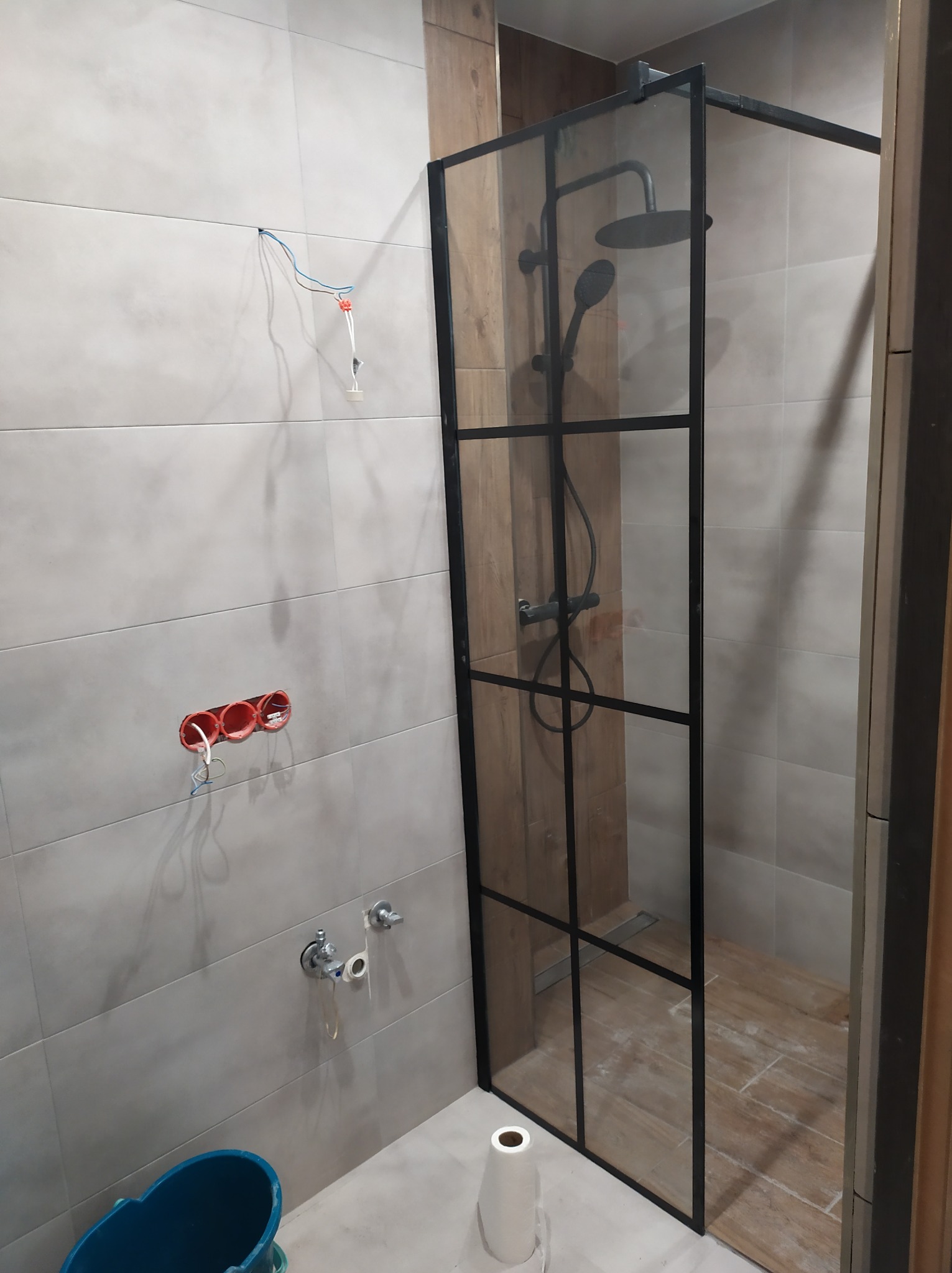 Montaż kabiny prysznicowej typu walk-in z czarnymi profilami, widoczne niedokończone podłączenia elektryczne i hydrauliczne na tle szarych płytek.