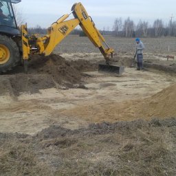 ZDAN -BUDOWNICTWO Sp. z o.o. - Żółta koparko-ładowarka JCB podczas prac ziemnych przy wykopie fundamentów, pracownik z łopatą wyrównuje teren, widoczne drewniane paliki wyznaczające obrys.