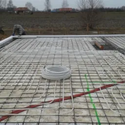 Przygotowanie płyty fundamentowej z instalacją ogrzewania podłogowego, widoczne zbrojenie, rury grzewcze w kolorze białym i czerwonym, oraz izolacja obwodowa z białego styropianu.