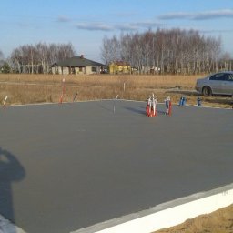 ZDAN -BUDOWNICTWO Sp. z o.o. - Wylana płyta fundamentowa z wystającymi rurami instalacyjnymi, zabezpieczona styropianem, z pracownikiem w tle i zaparkowanym samochodem osobowym.
