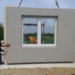 ZDAN -BUDOWNICTWO Sp. z o.o. - Montaż prefabrykowanej ściany z oknem na fundamencie. Pracownik przytrzymuje ścianę, widoczny dźwig i odbicie drugiego pracownika w oknie.