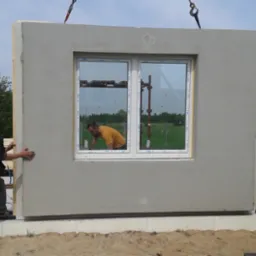 Montaż prefabrykowanej ściany z oknem na fundamencie. Pracownik przytrzymuje ścianę, widoczny dźwig i odbicie drugiego pracownika w oknie.