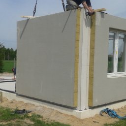 ZDAN -BUDOWNICTWO Sp. z o.o. - Montaż prefabrykowanych ścian domu na fundamencie, widoczne ocieplenie między panelami, okno z szybami zespolonymi, pracownik na dachu oraz narzędzia na płycie fundamentowej.