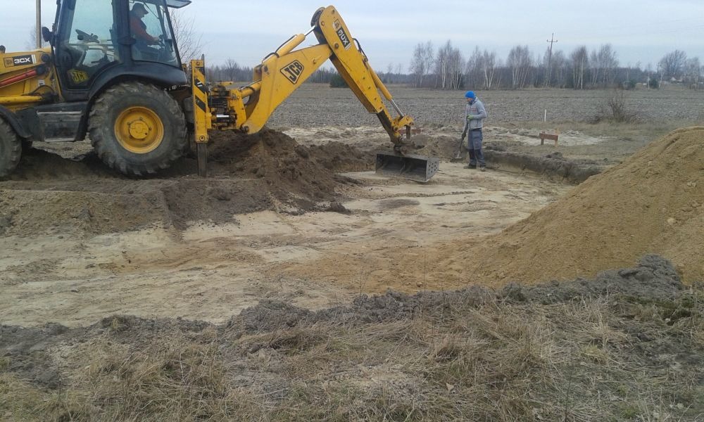 Żółta koparko-ładowarka JCB podczas prac ziemnych przy wykopie fundamentów, pracownik z łopatą wyrównuje teren, widoczne drewniane paliki wyznaczające obrys.
