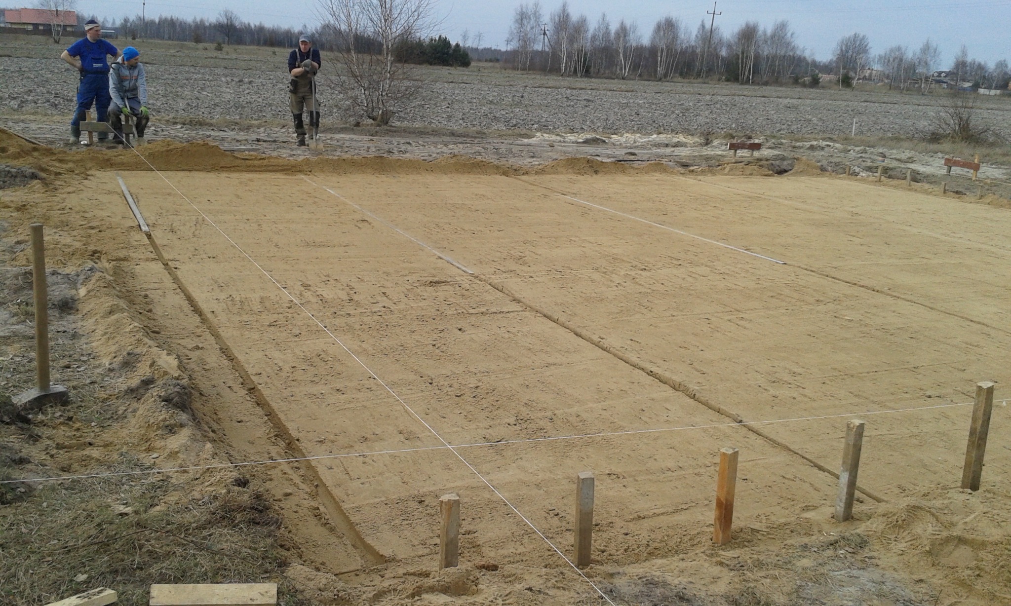 Przygotowany teren pod fundamenty domu z wytyczonymi liniami i wbitymi słupkami, widoczni pracownicy w tle.