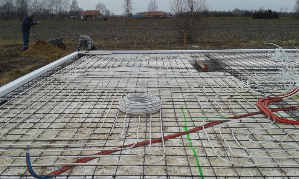Przygotowanie płyty fundamentowej z instalacją ogrzewania podłogowego, widoczne zbrojenie, rury grzewcze w kolorze białym i czerwonym, oraz izolacja obwodowa z białego styropianu.
