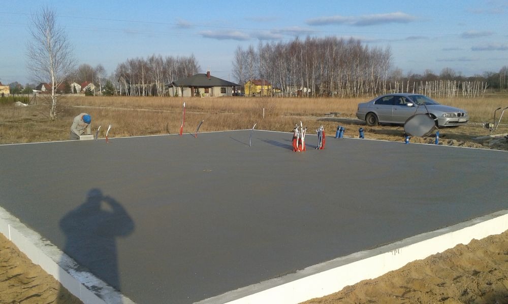 Wylana płyta fundamentowa z wystającymi rurami instalacyjnymi, zabezpieczona styropianem, z pracownikiem w tle i zaparkowanym samochodem osobowym.