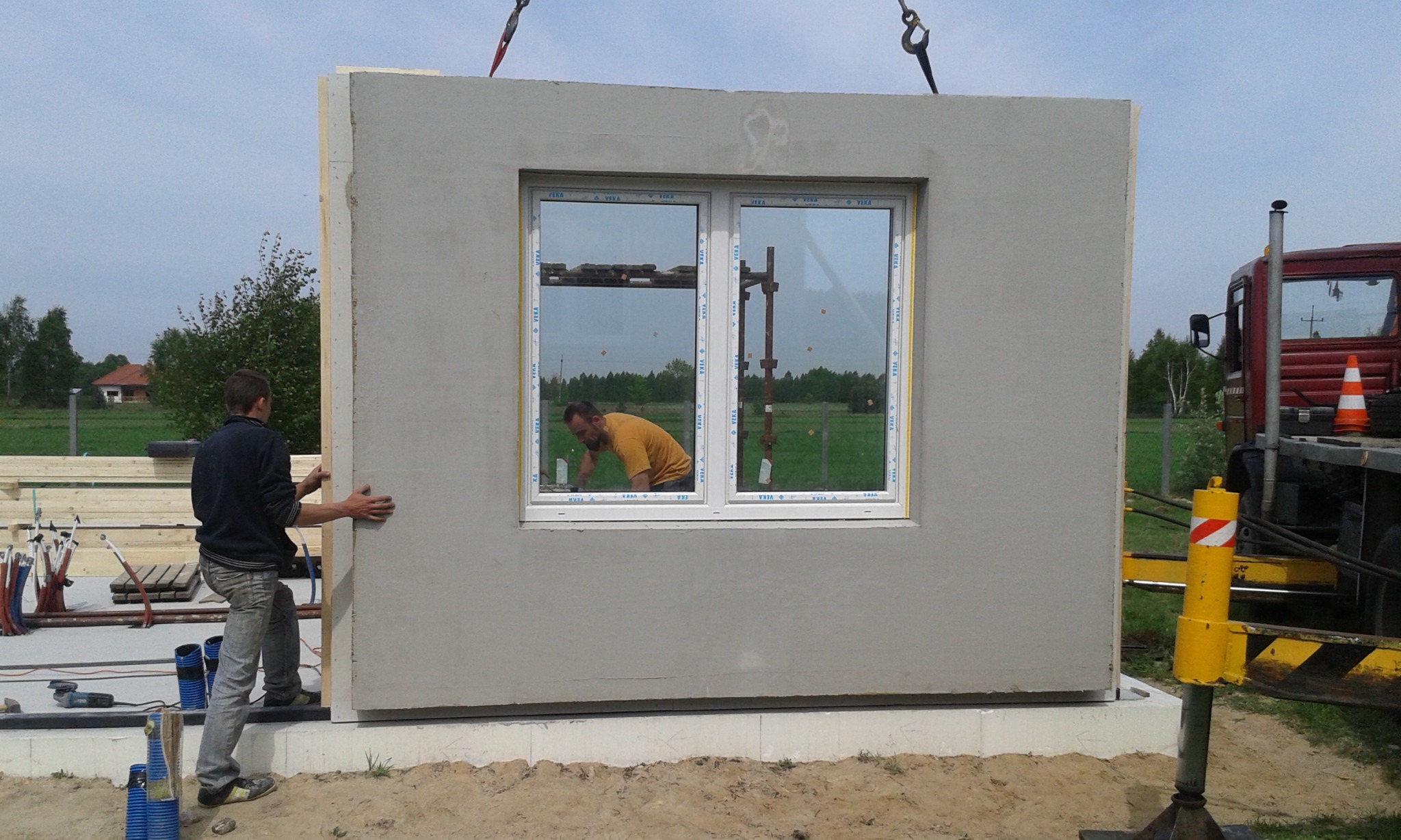 Montaż prefabrykowanej ściany z oknem na fundamencie. Pracownik przytrzymuje ścianę, widoczny dźwig i odbicie drugiego pracownika w oknie.