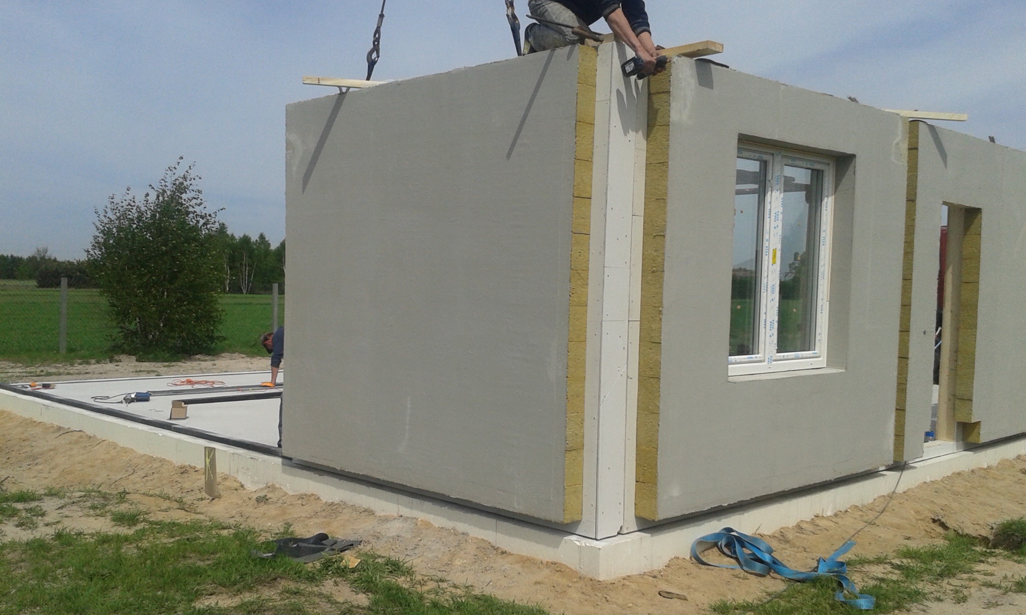 Montaż prefabrykowanych ścian domu na fundamencie, widoczne ocieplenie między panelami, okno z szybami zespolonymi, pracownik na dachu oraz narzędzia na płycie fundamentowej.
