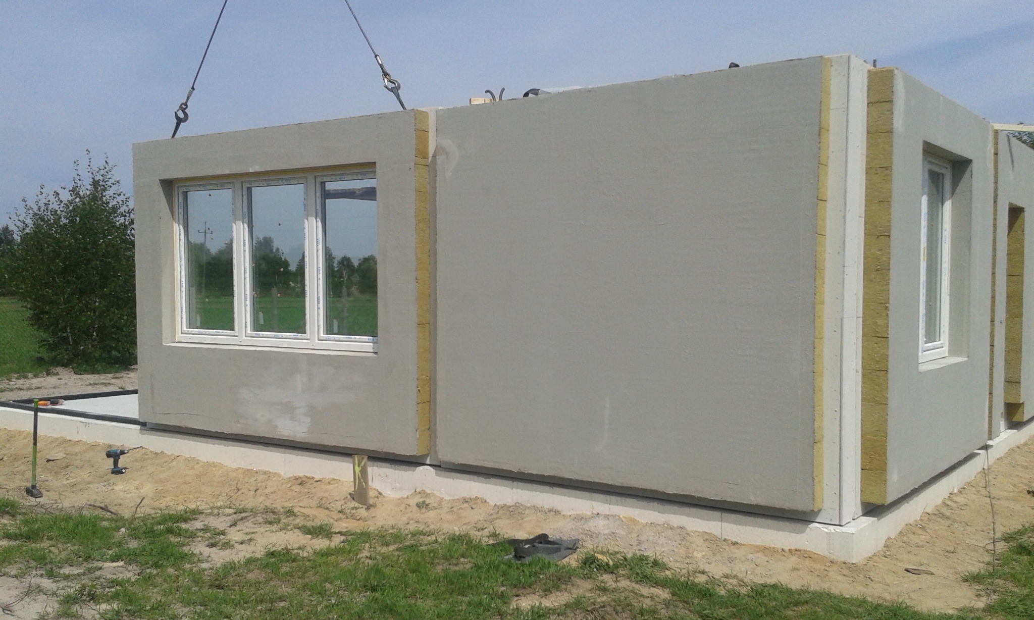 Montaż segmentu domu modułowego z oknem, widoczne elementy izolacji i liny dźwigu, w tle zieleń